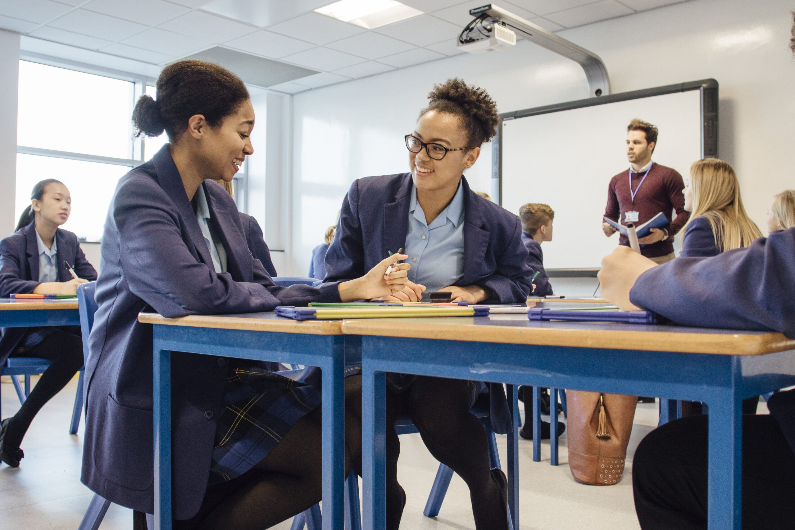 Two teenage girls in school uniforms in a classroom talking